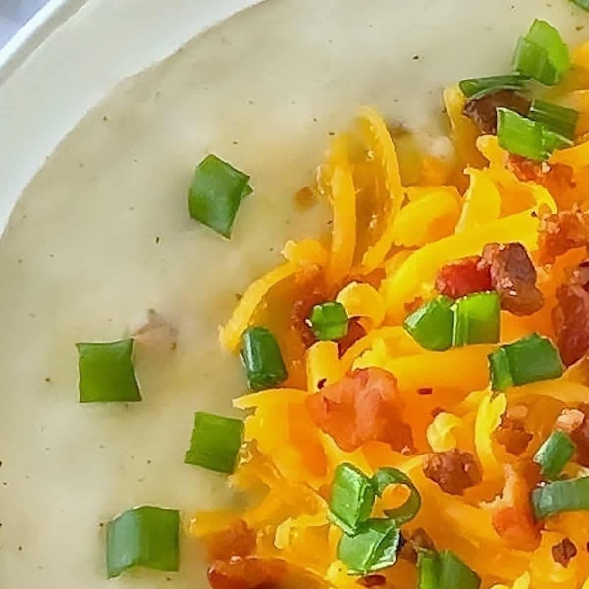 Close up of potato soup in a white bowl with cheese, bacon, and green onions on top.