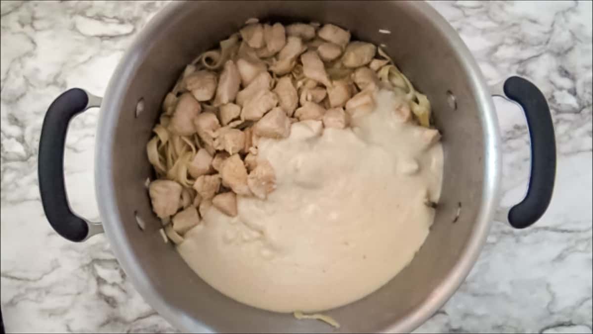 Alfredo sauce poured into a large pot containing cooked fettuccine pasta and cubed chicken.