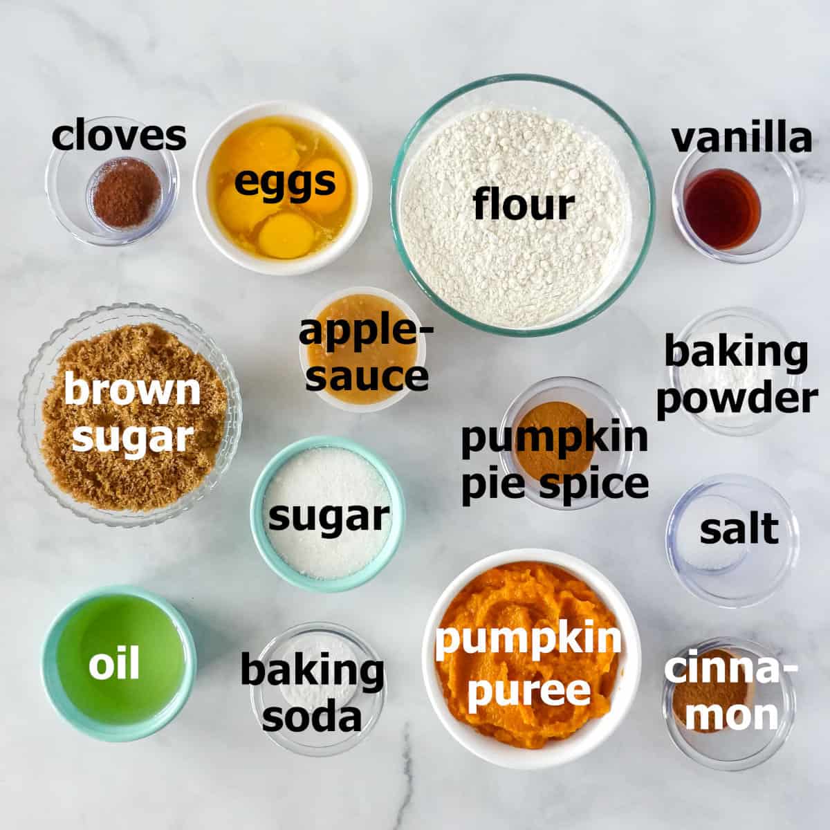 Ingredients for pumpkin cupcakes in separate bowls on a white marble countertop.