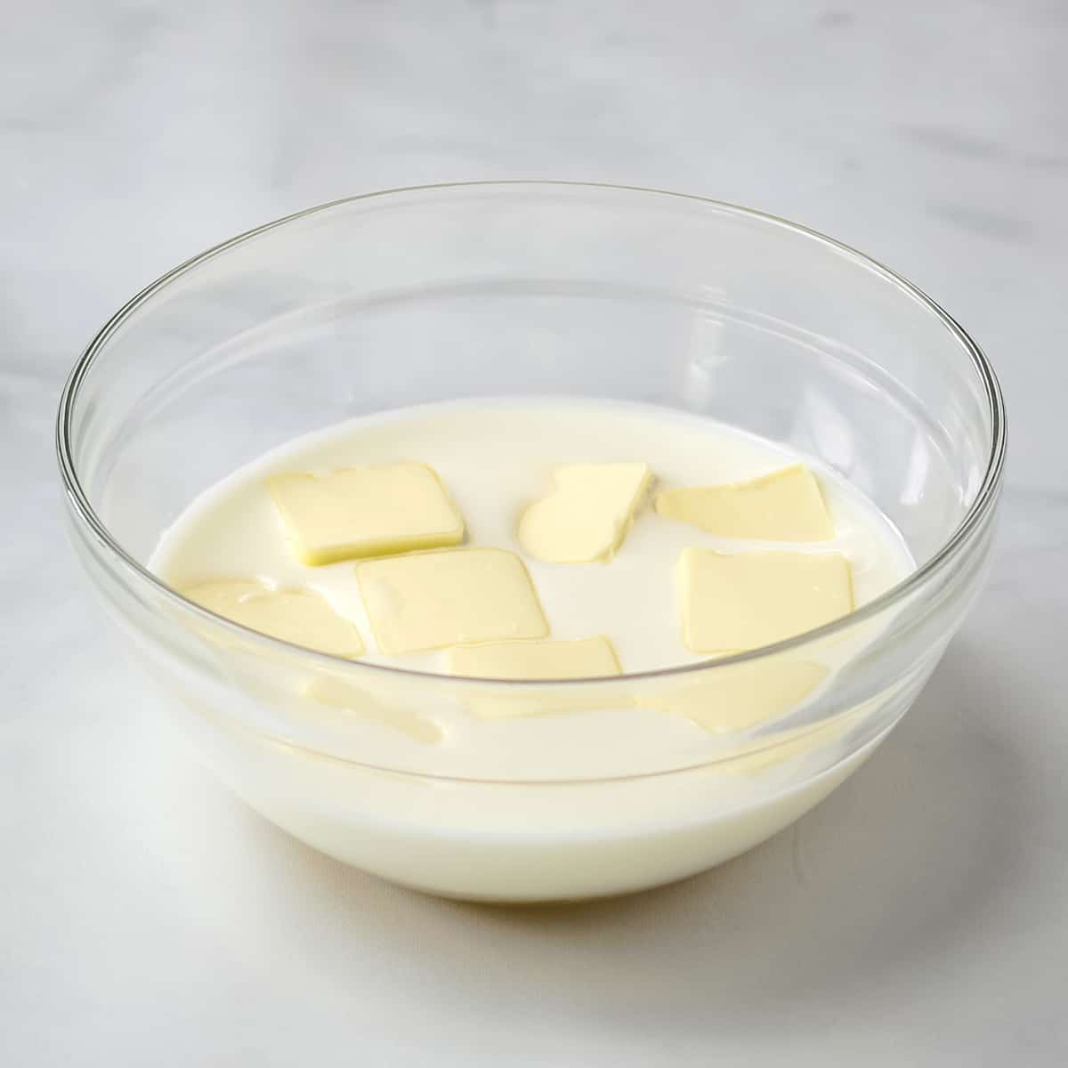 Sliced butter pads in a glass bowl filled with milk and sugar.