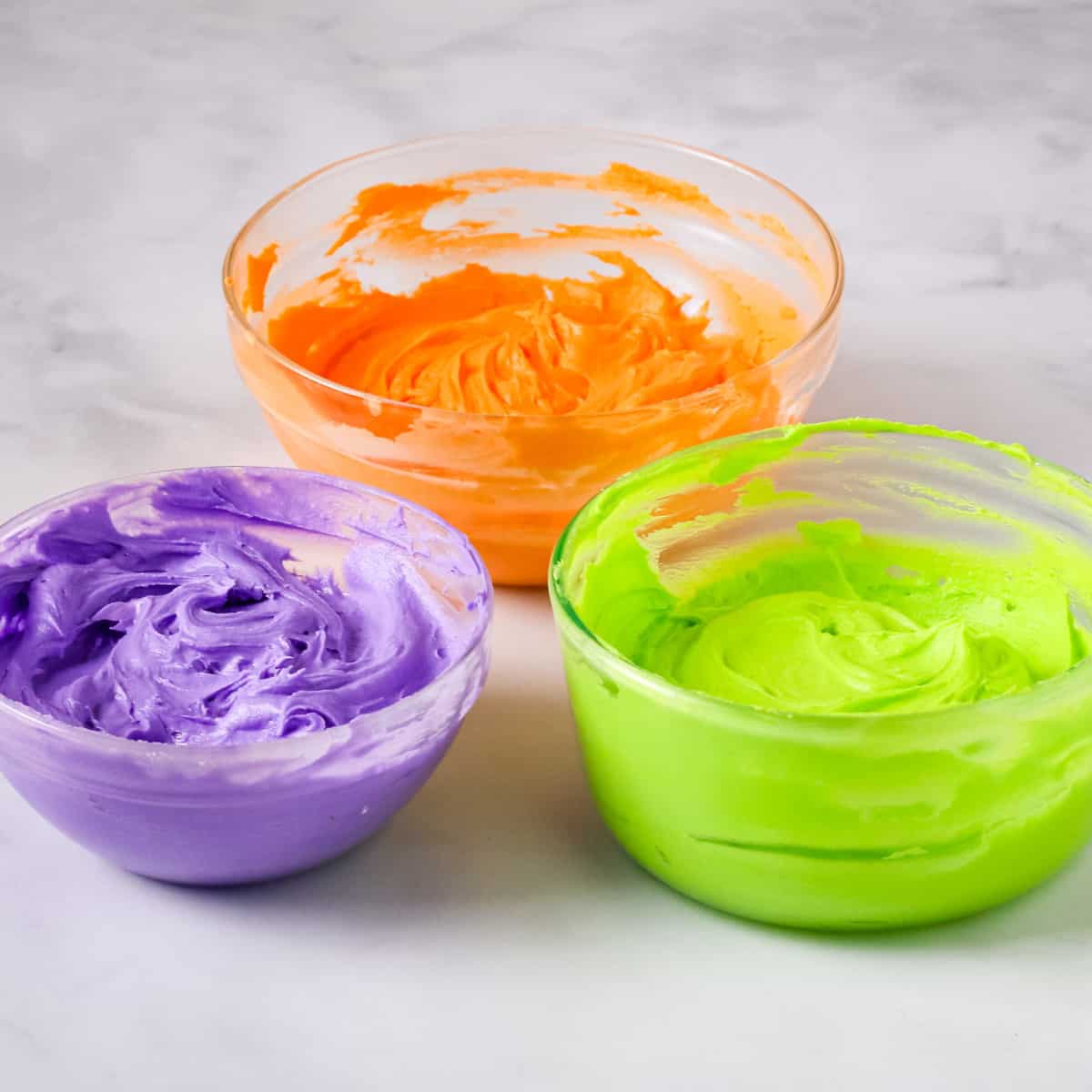 Purple, orange, and green colored buttercream in 3 separate glass bowls.