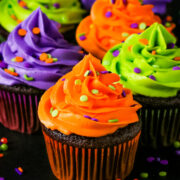 Halloween themed chocolate cupcakes topped with neon green, orange, and purple colored buttercream with sprinkles on a black table.