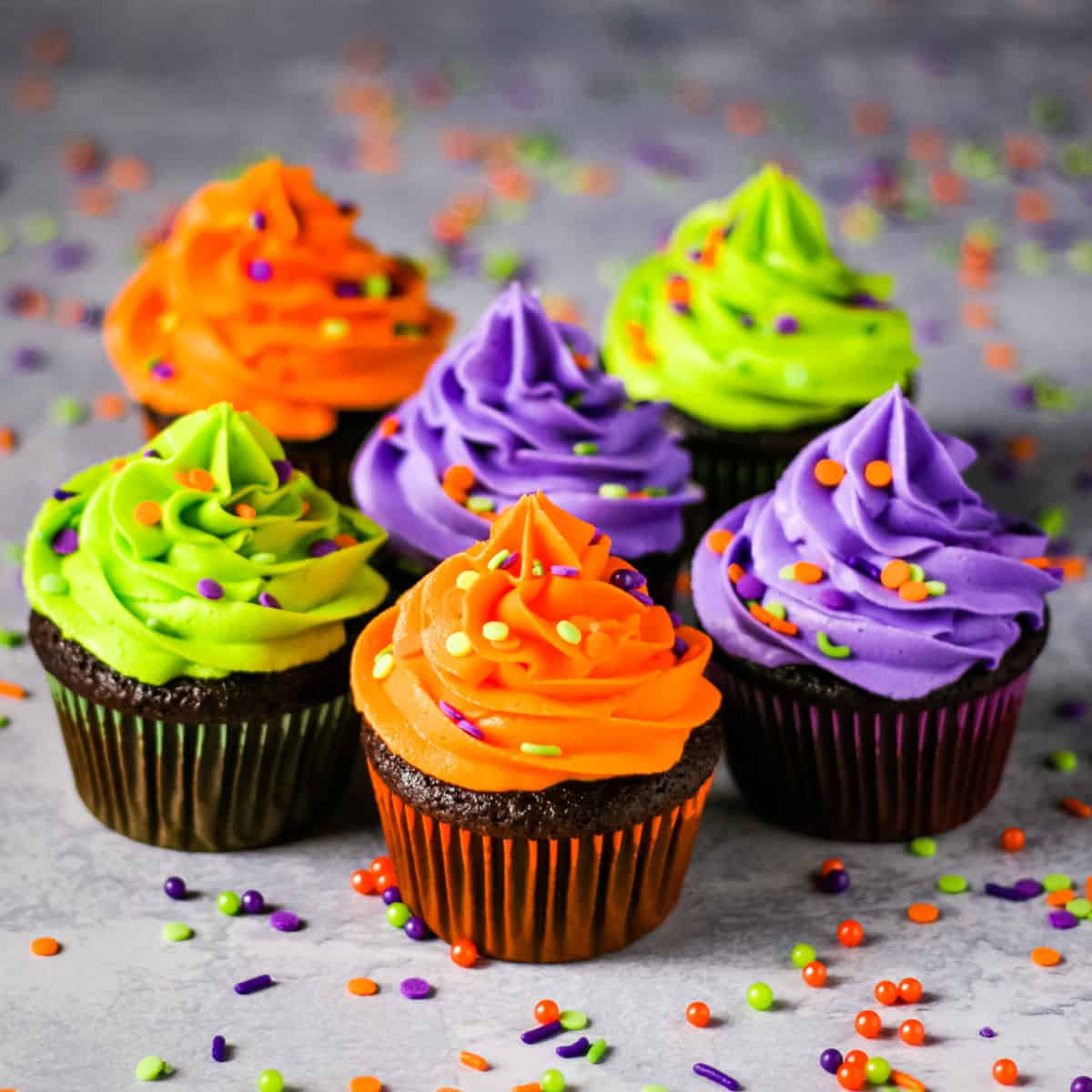 Halloween themed chocolate cupcakes topped with neon green, orange, and purple colored buttercream with sprinkles on a concrete table.