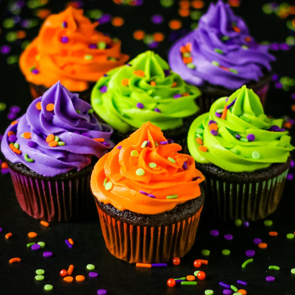 Halloween themed chocolate cupcakes topped with neon green, orange, and purple colored buttercream with sprinkles on a black table.