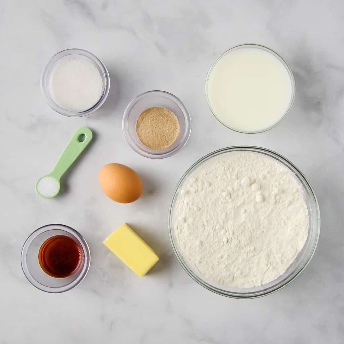 Cinnamon roll ingredients on a white marble countertop: flour, buttermilk, butter, sugar, yeast, egg, vanilla extract, and salt.