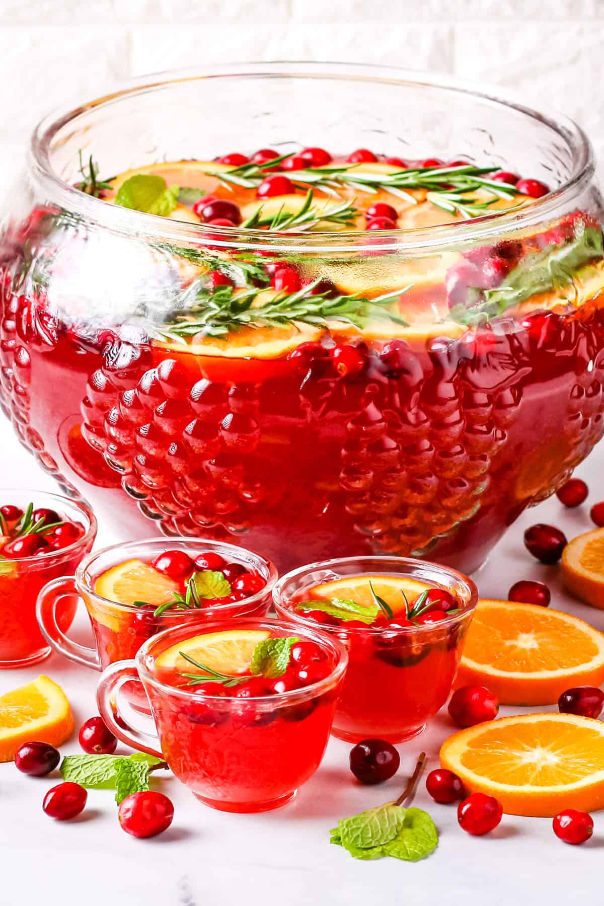 Christmas punch in a large glass punch bowl and in small glass cups garnished with orange slices, cranberries, rosemary sprigs, and mint leaves.
