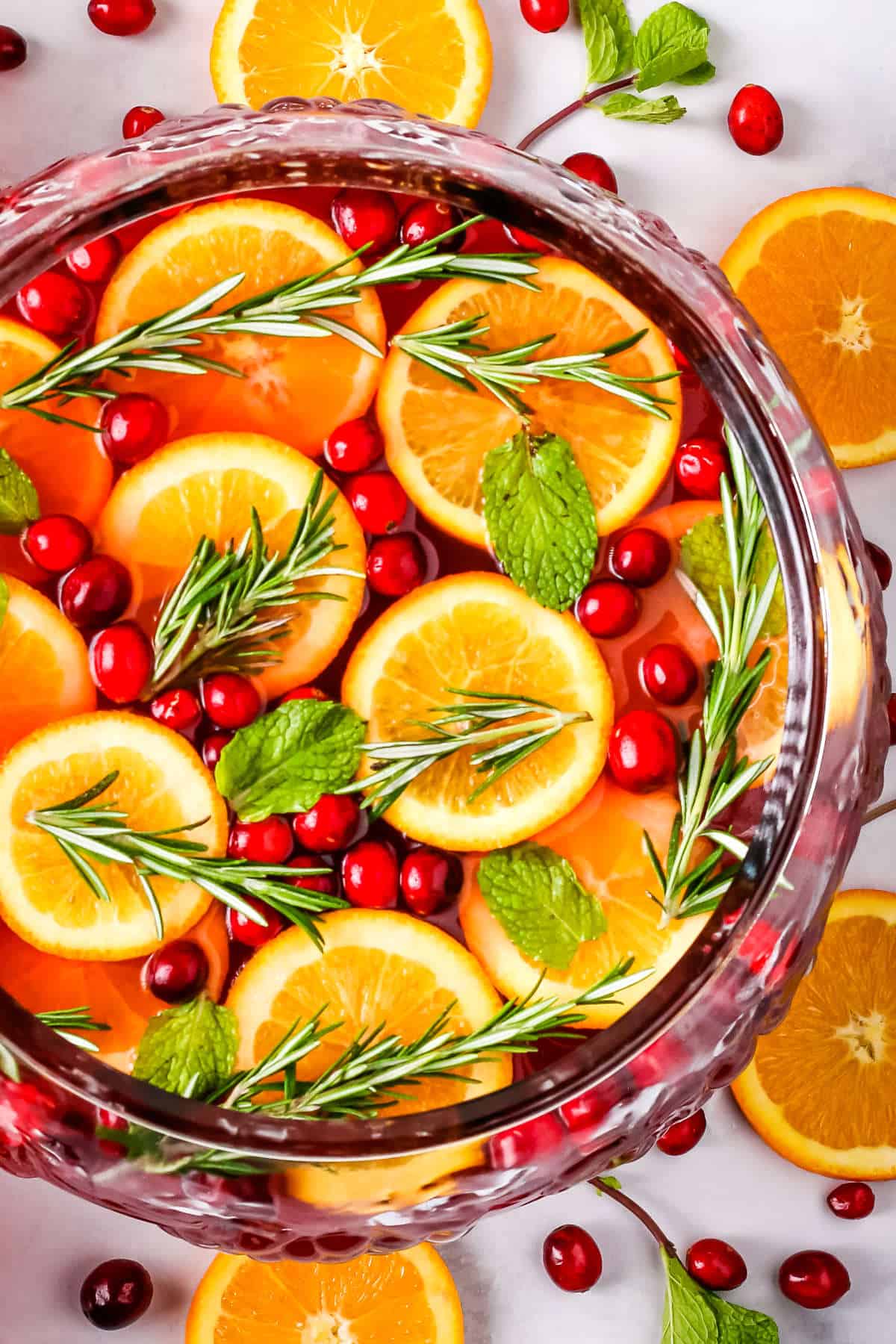 Christmas punch in a large glass punch bowl garnished with orange slices, cranberries, rosemary sprigs, and mint leaves.