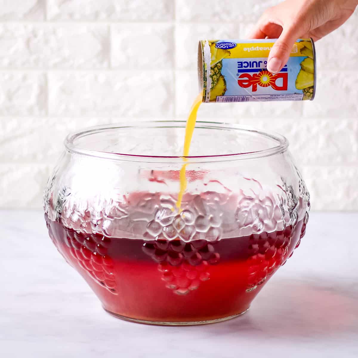 Pouring pineapple juice into a large glass punch bowl.