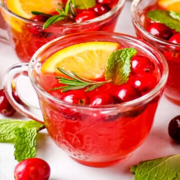 Christmas punch in a small glass cup garnished with an orange slice, cranberries, rosemary sprig, and mint leaf.