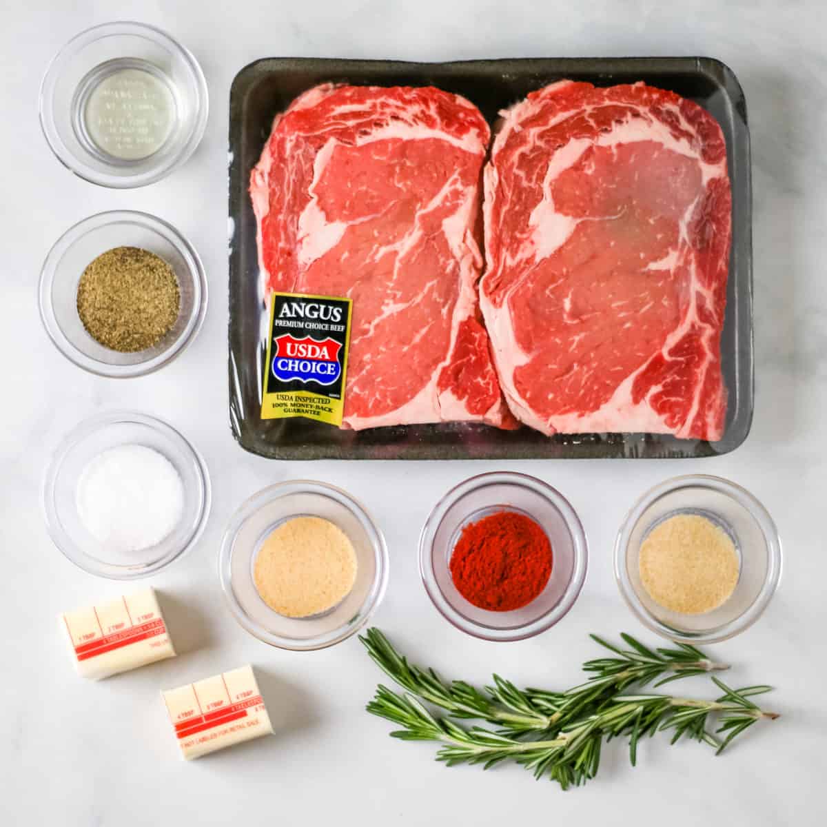 Ingredients for pan seared steak: ribeye steak, black pepper, salt, paprika, garlic powder, onion powder, canola oil, butter, and fresh rosemary.