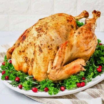 A front view of a whole turkey that has been baked in an oven bag on a white serving dish garnished with grape tomatoes and herbs.