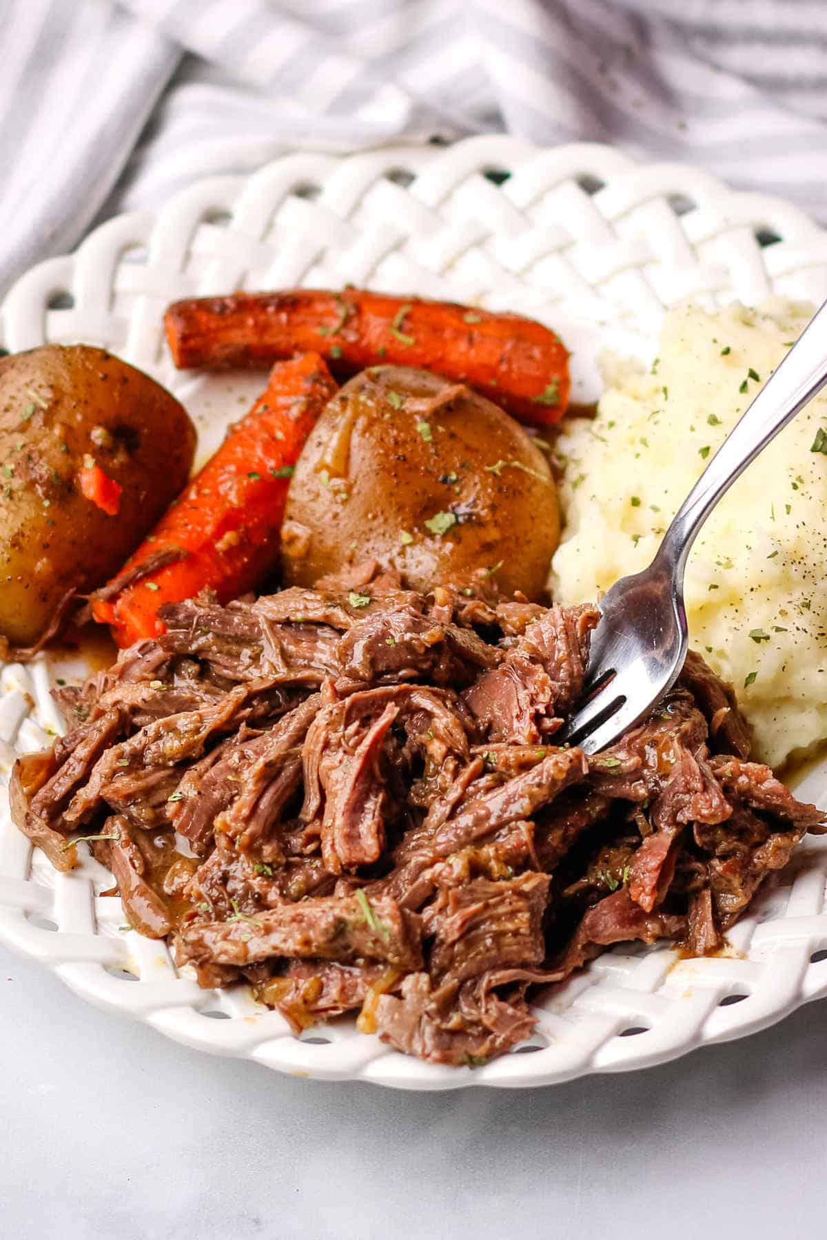 Slow cooker venison roast recipe served on a white plate with carrots and potatoes.