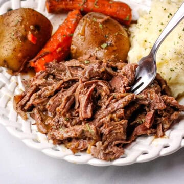 Slow cooker venison roast recipe served on a white plate with carrots and potatoes.