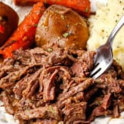 Slow cooker venison roast recipe served on a white plate with cooked carrots and potatoes.